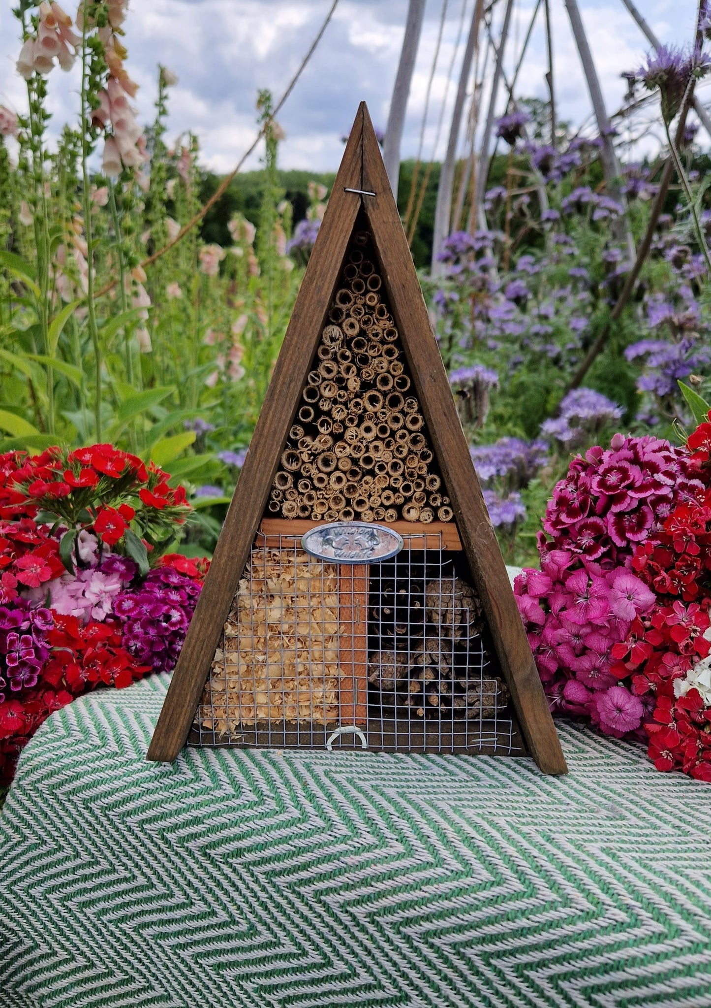 A triangular shaped wooden structure, filled with different textures of wood, on top of a green blanket surrounded by flowers in the walled garden at harewood.