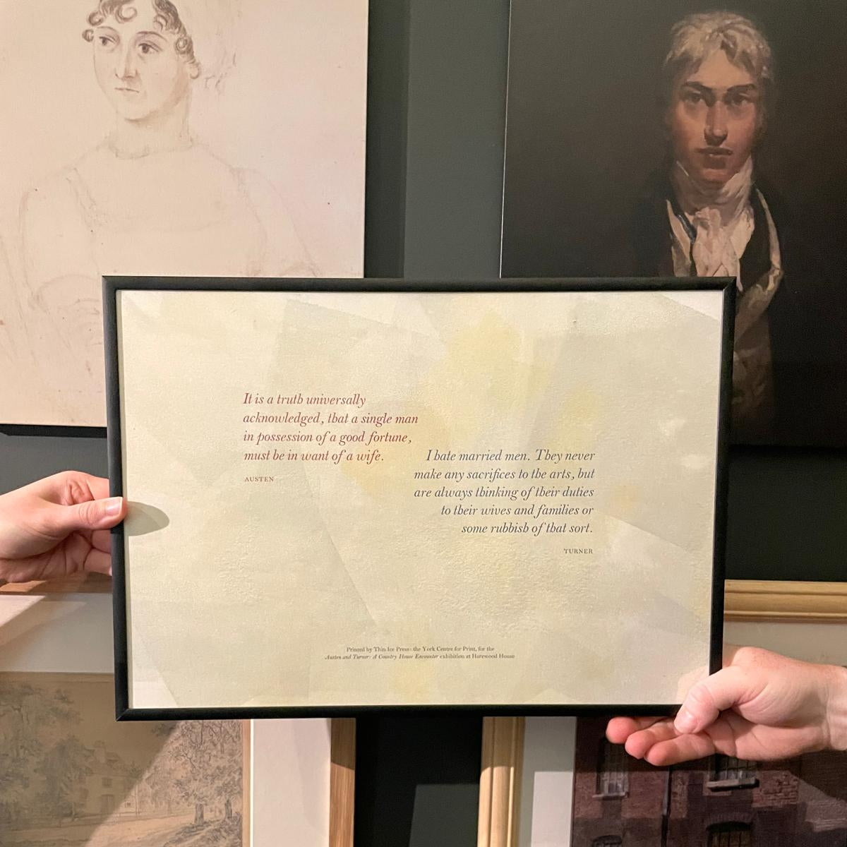Person holding a framed document with text against a wall with framed artworks.