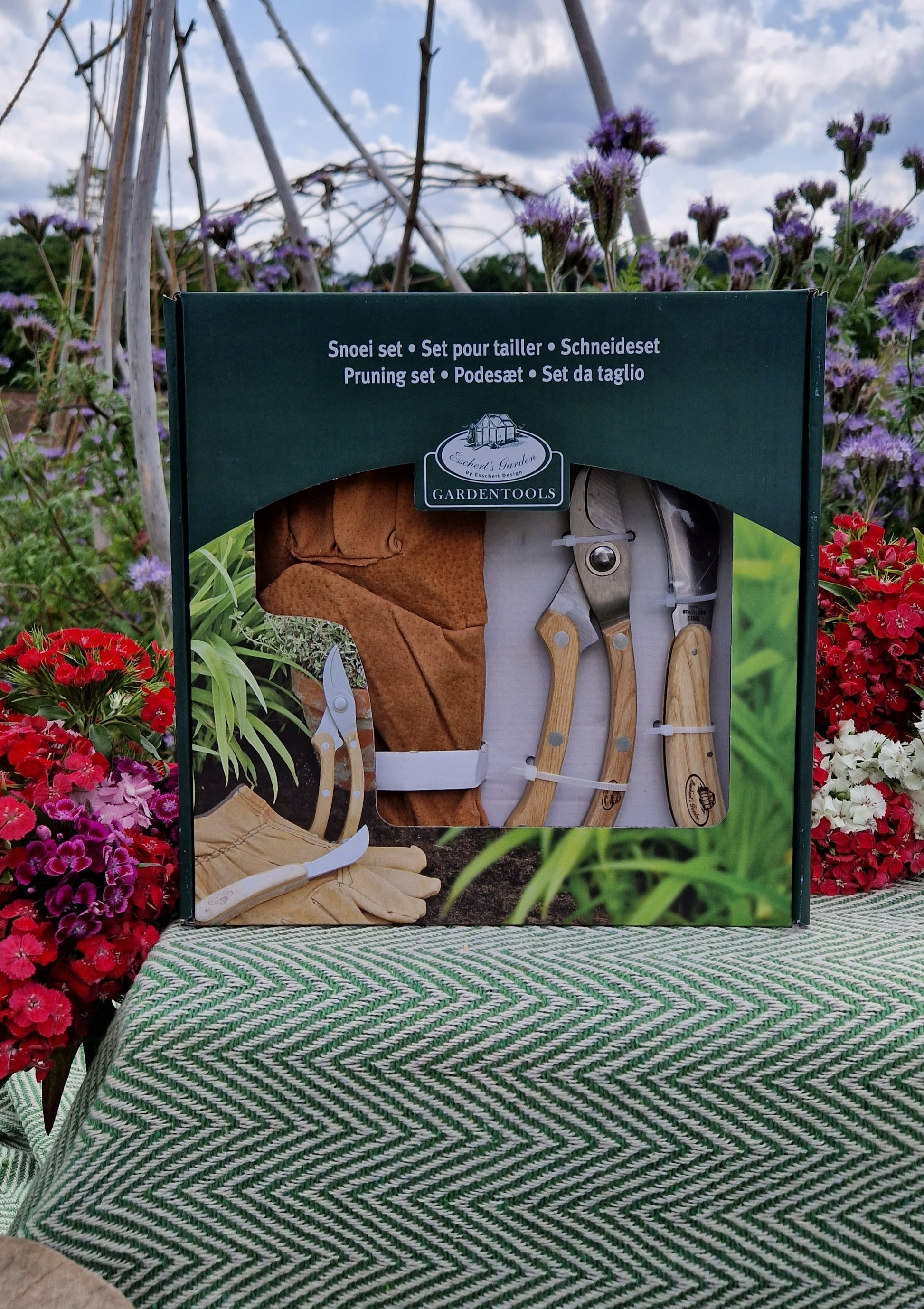 A garden pruning set containing sheers, a knife and a pair of brown leather gloves in a green box, propped against a crate covered in a green blanket in a garden.